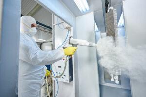 Worker in protective gear spray-coating a metal part in an industrial facility.
