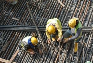 Three construction workers in helmets and reflective vests tying rebar at a construction site.