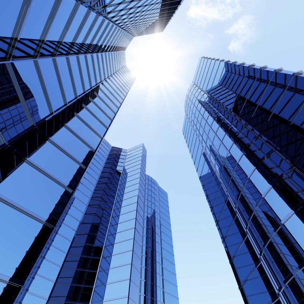 Tall glass skyscrapers with reflective windows viewed from below toward the sun and blue sky.