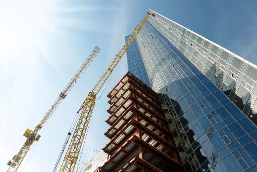 Steel building under construction beside a glass skyscraper with two yellow cranes, daytime.