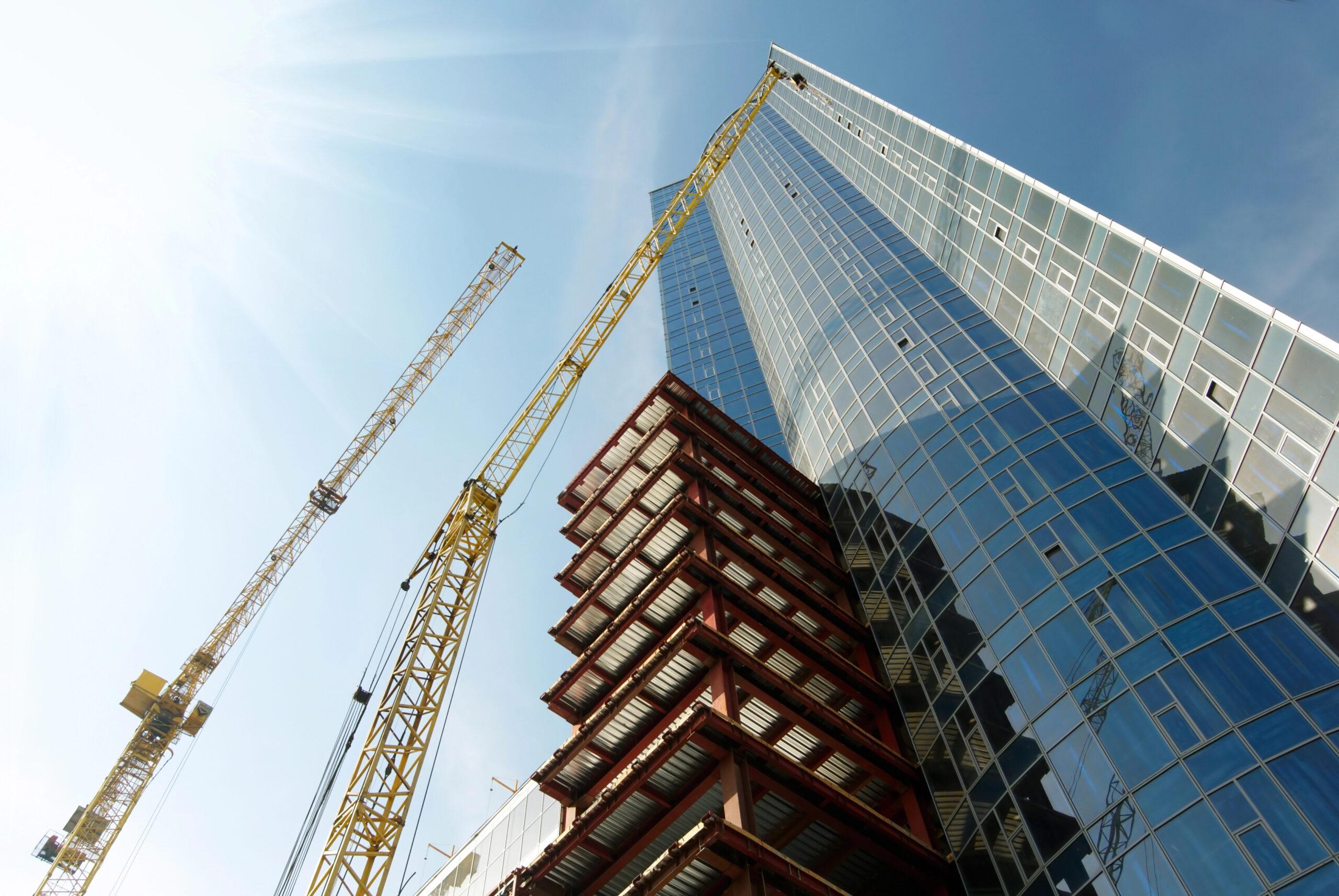 Steel building under construction beside a glass skyscraper with two yellow cranes, daytime.