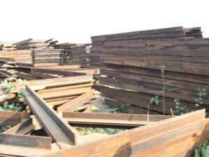 Stacks of rusty steel beams and metal sheets are arranged outdoors on bare ground with some plants.