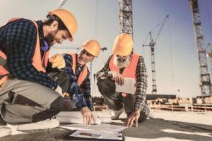 Three construction workers in safety gear review blueprints at a building site with cranes.
